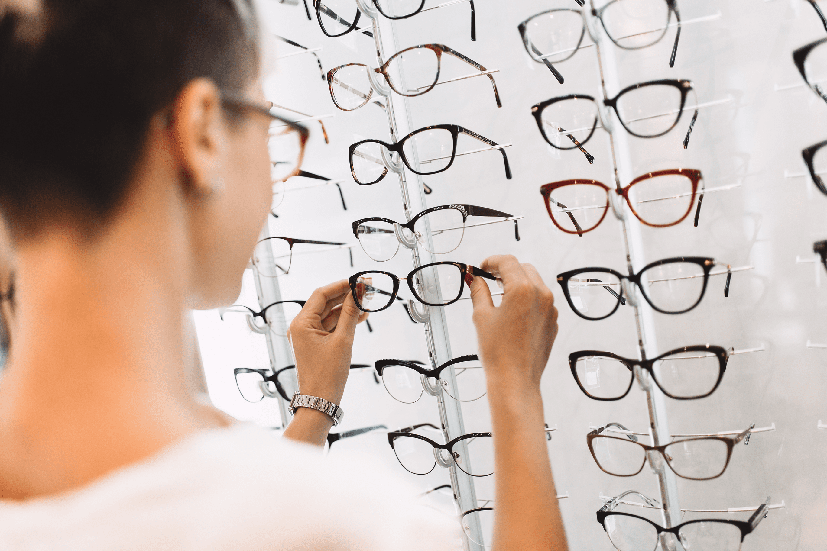 A woman holds up a pair of glasses in a shop in front of a mirror